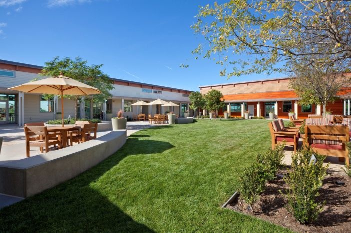 View of the OASIS Senior Center courtyard.  Large grass space surrounded by concrete and patio tables with umbrellas and chairs.