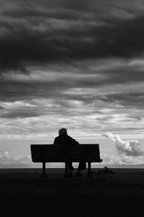 Photograph of a silhouetted person sitting on a bench with clouds in the sky.