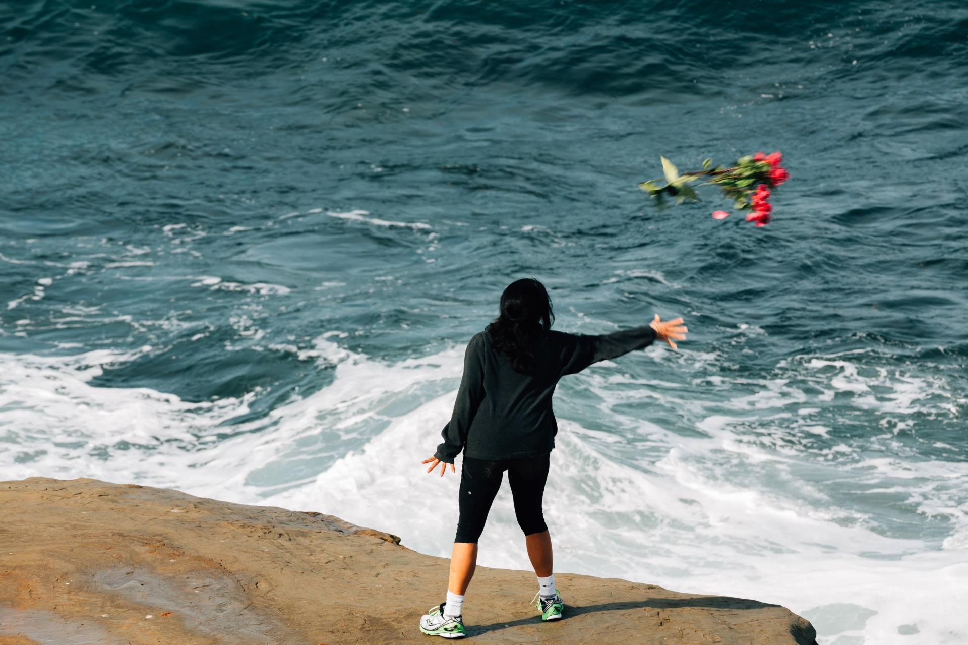 Photography of a woman standing at the edge of the water tossing flowers into the sea.